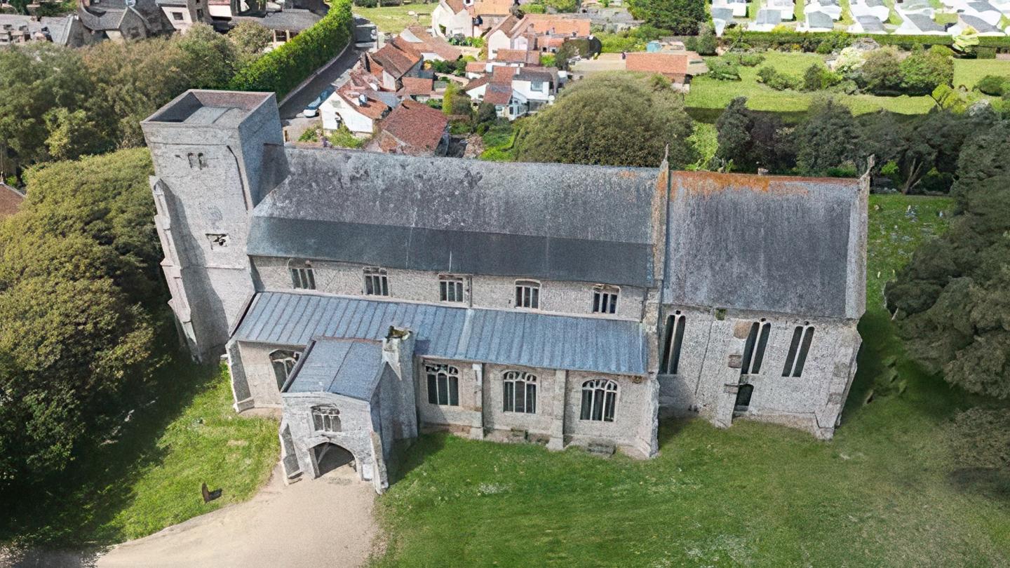 An aerial view of All Saints Church, Thornham - a large stone church with a square tower and south porch, seen from above among its churchyard and village rooftops
